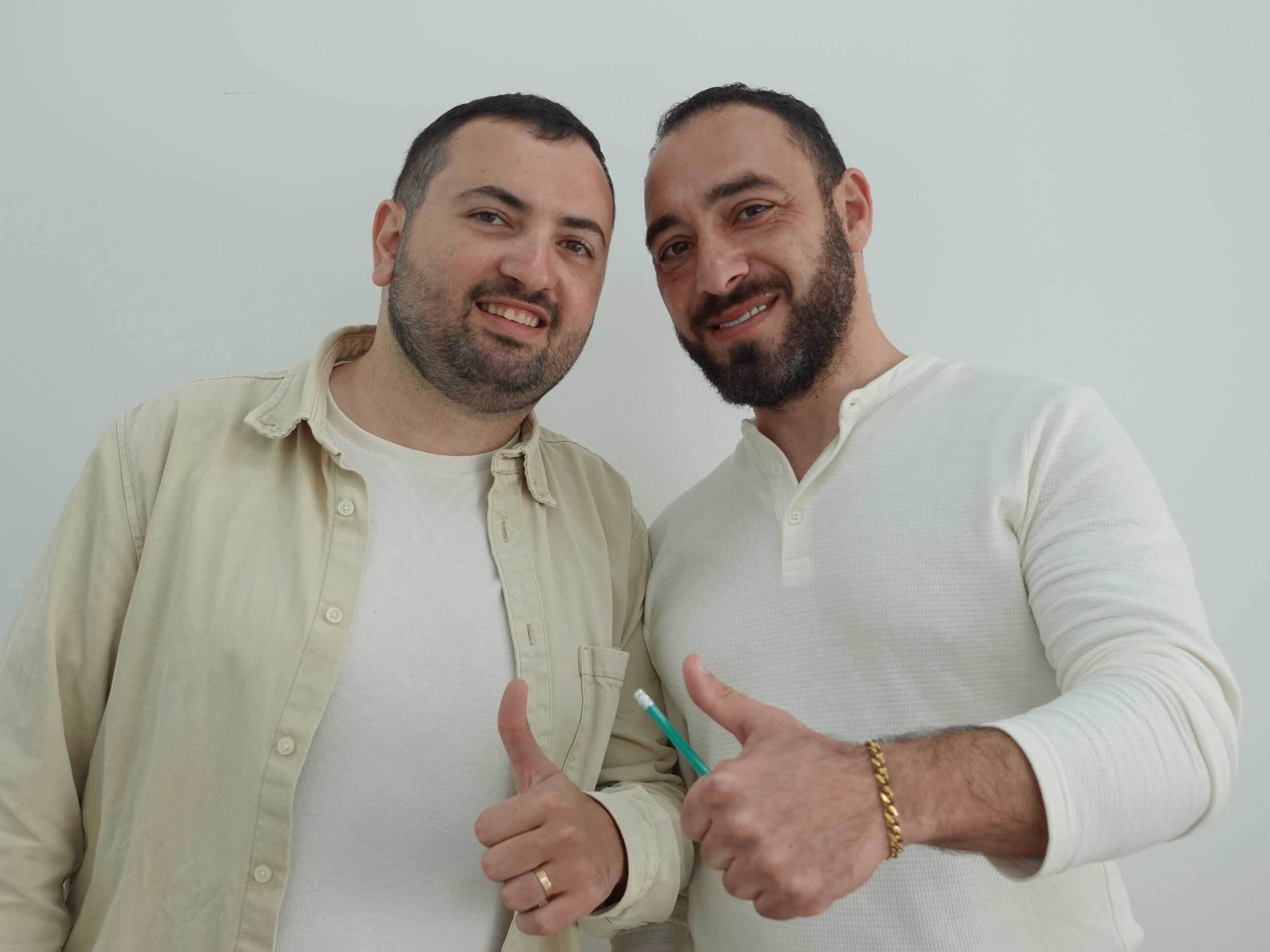 שני גברים מזוקנים מחייכים עם אגודלים מורמים, אחד מחזיק עיפרון.
